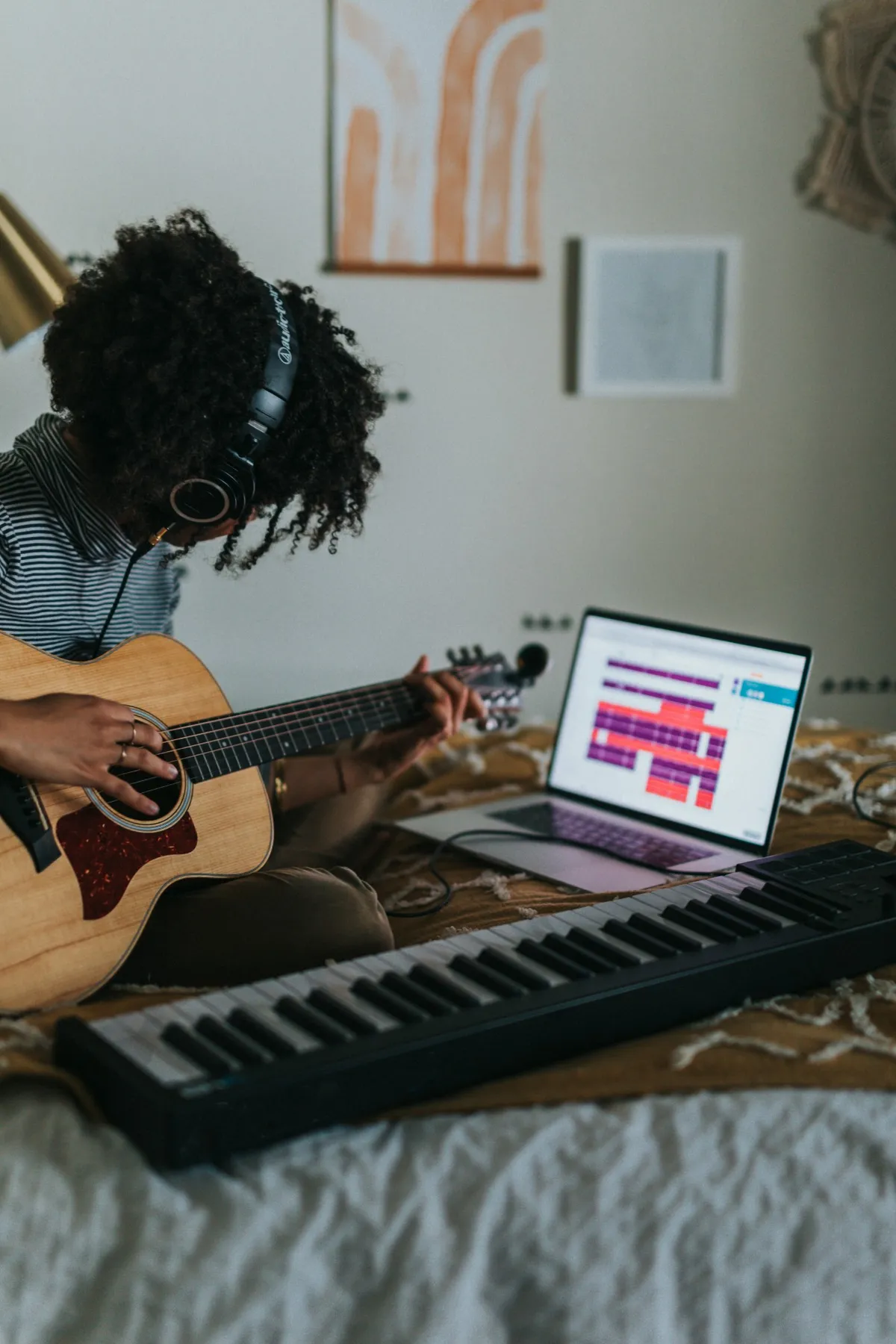 A musician in headphones playing acoustic guitar beside a laptop and keyboard, recording at home